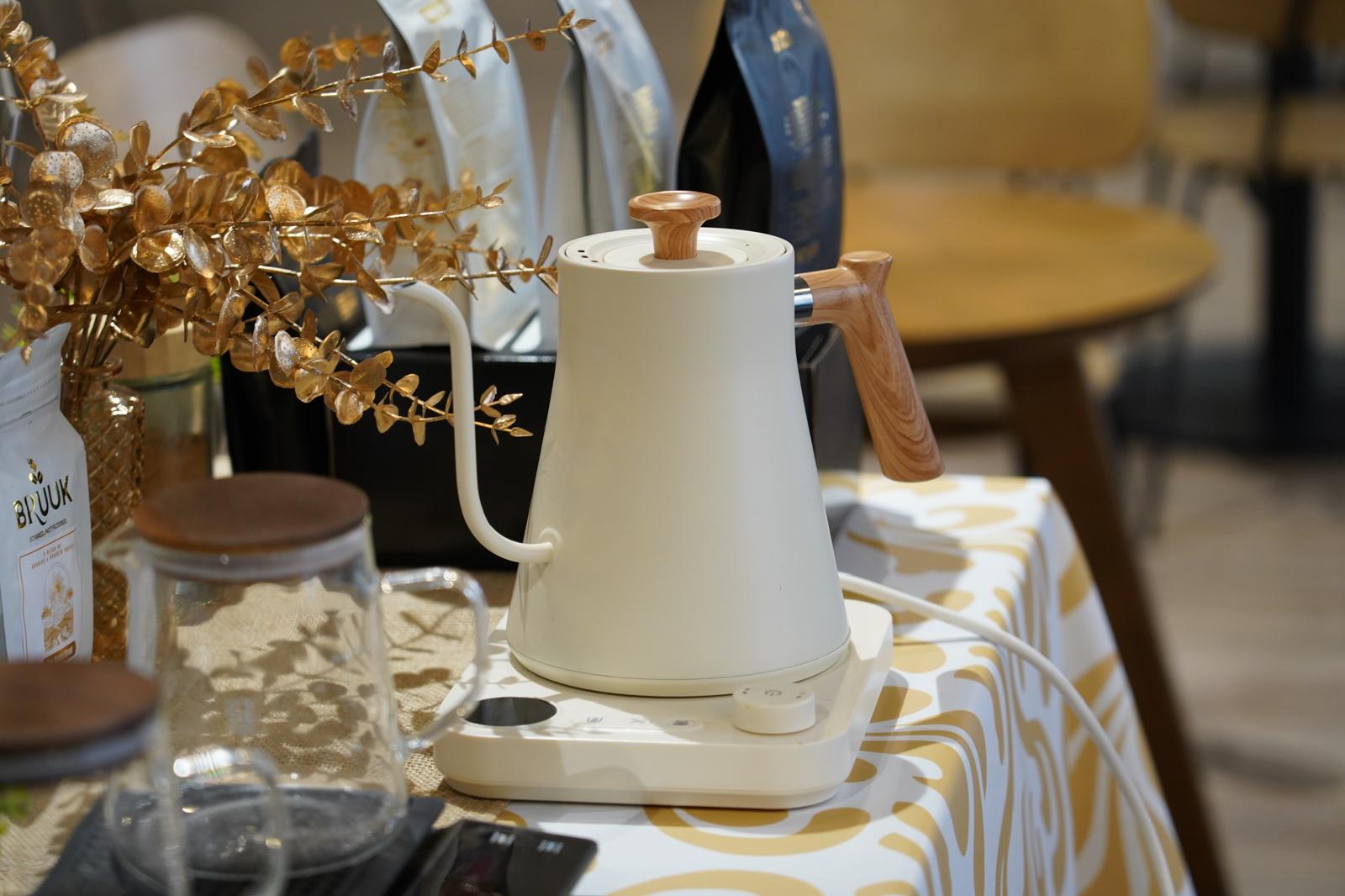 A cream gooseneck kettle on a coffee bar, with BRUUK coffee bags and glass carafes around it